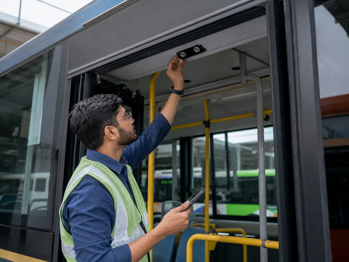 An Indian field engineer installing and calibrating a passenger-counting sensor above a city bus doorway